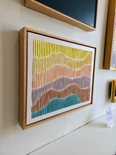 Framed abstract artwork featuring wavy, colorful stripes in yellow, pink, gray, and blue tones.