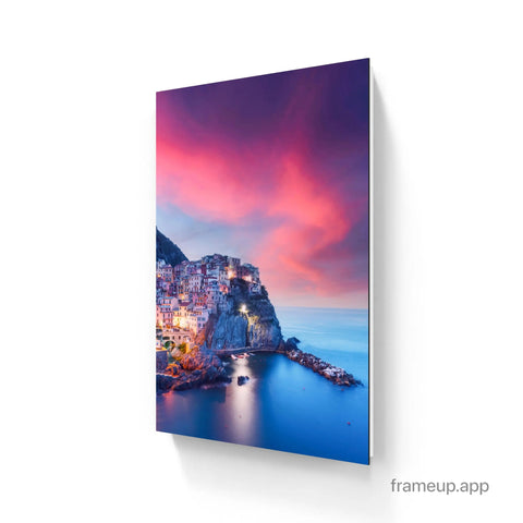 A vibrant canvas print depicting a cliffside coastal village at twilight, showcasing colorful buildings against a dramatic pink and purple sky reflected in the calm blue water.