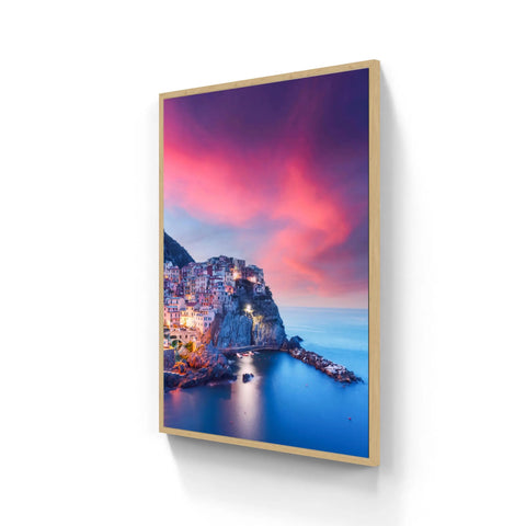 A rectangular framed canvas print featuring a vibrant coastal village at dusk, with warm-toned buildings nestled on cliffs against a dramatic pink and purple sky over calm blue water.