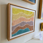 Framed abstract artwork featuring wavy, colorful stripes in yellow, pink, gray, and blue tones.