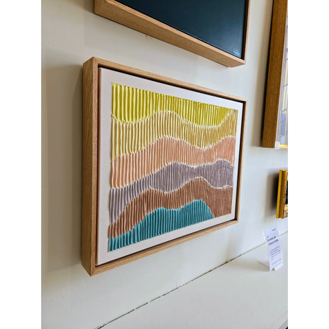 Framed abstract artwork featuring wavy, colorful stripes in yellow, pink, gray, and blue tones.