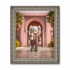 A kaws-style beige figurine with gray cloud hair and black x eyes holding an ice cream in front of pink moroccan architecture in original aluminium artwork